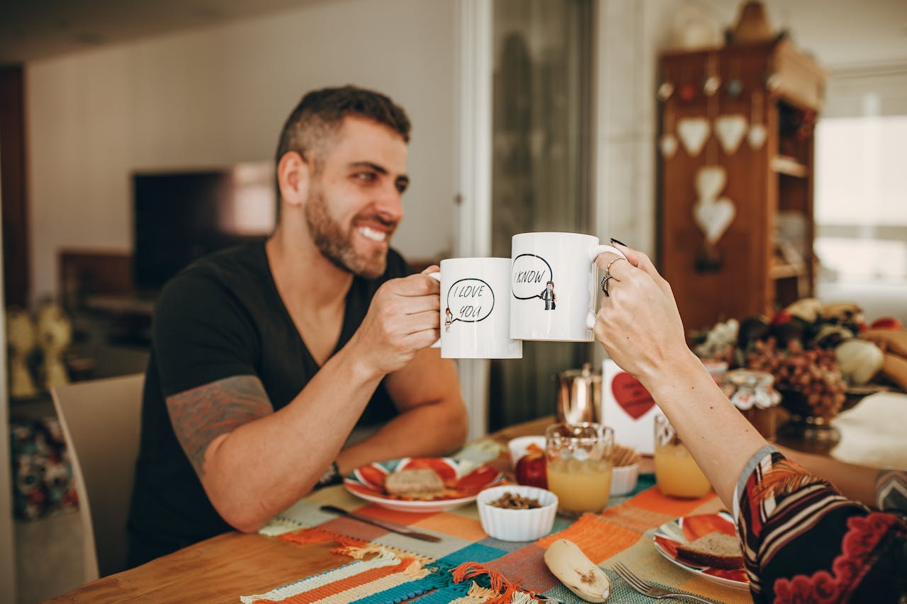 hero-img-01 A joyful couple toasting with customized mugs during breakfast at home.