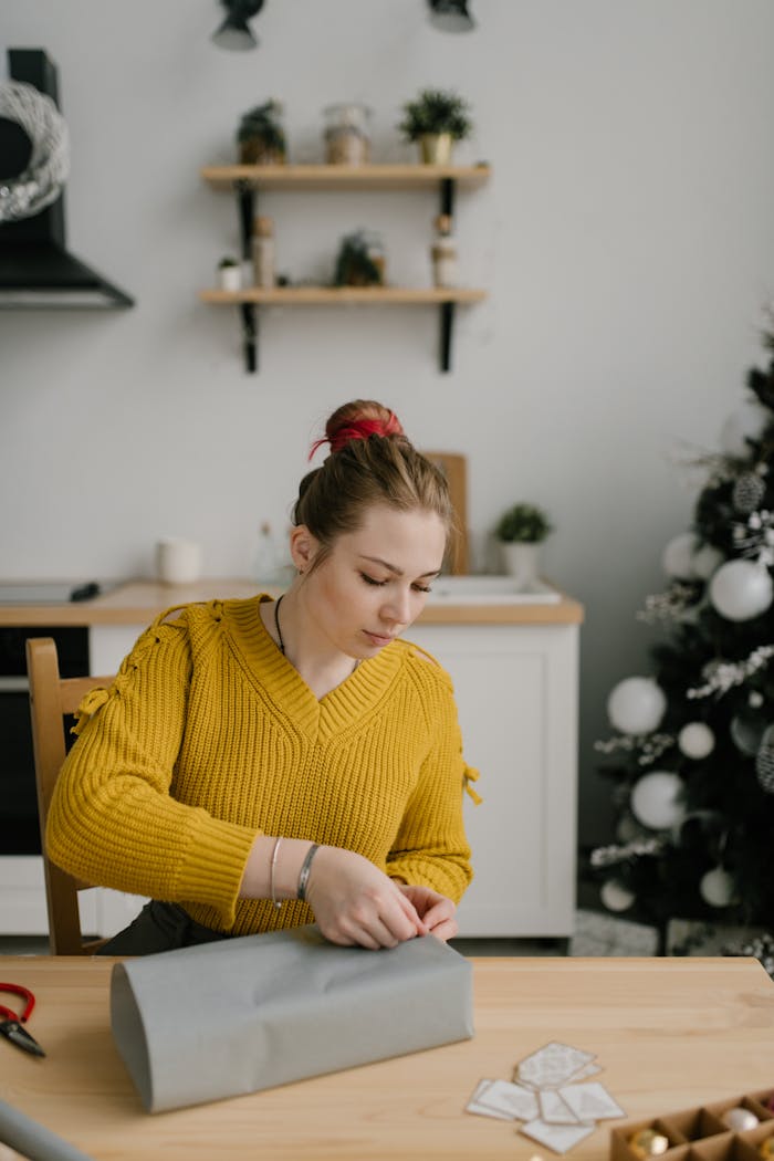 why-choose-us A woman in a cozy sweater wraps gifts at a table with a Christmas tree in the background.