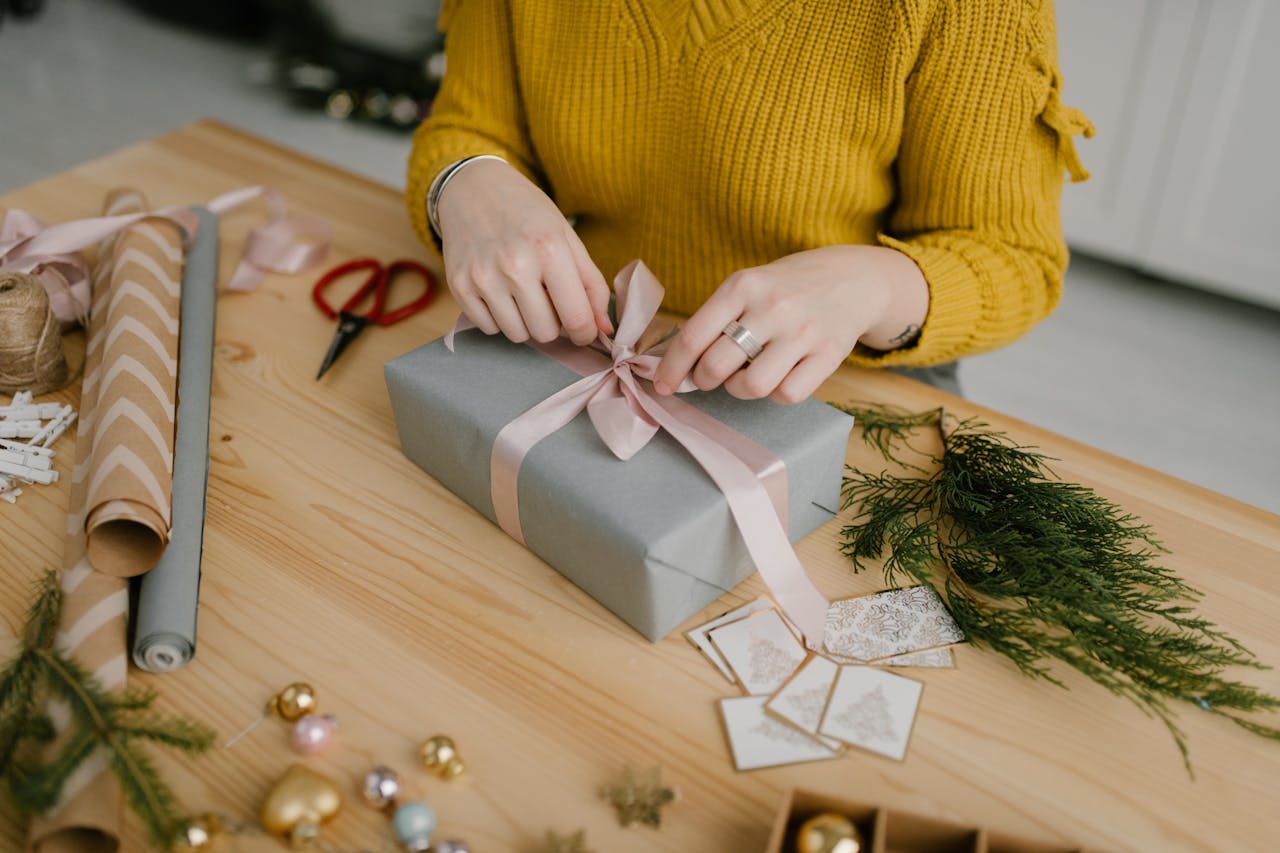 hero-img-02 Woman in yellow sweater wrapping a gift with pink ribbon on wooden table decorated for Christmas.