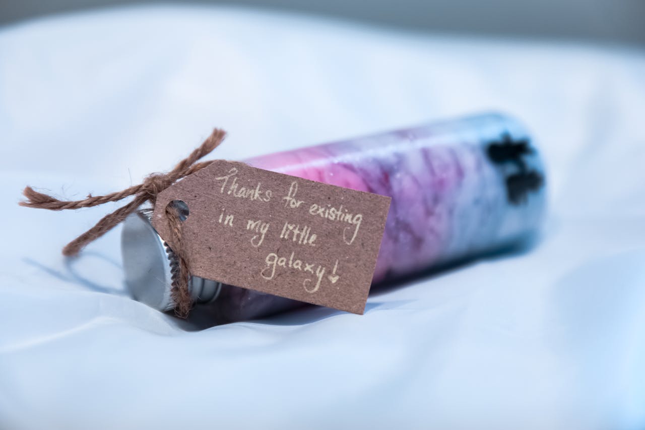 gallery-2 A candle with purple hues and a heartfelt message on a kraft tag, saying 'Thanks for existing in my little galaxy'.